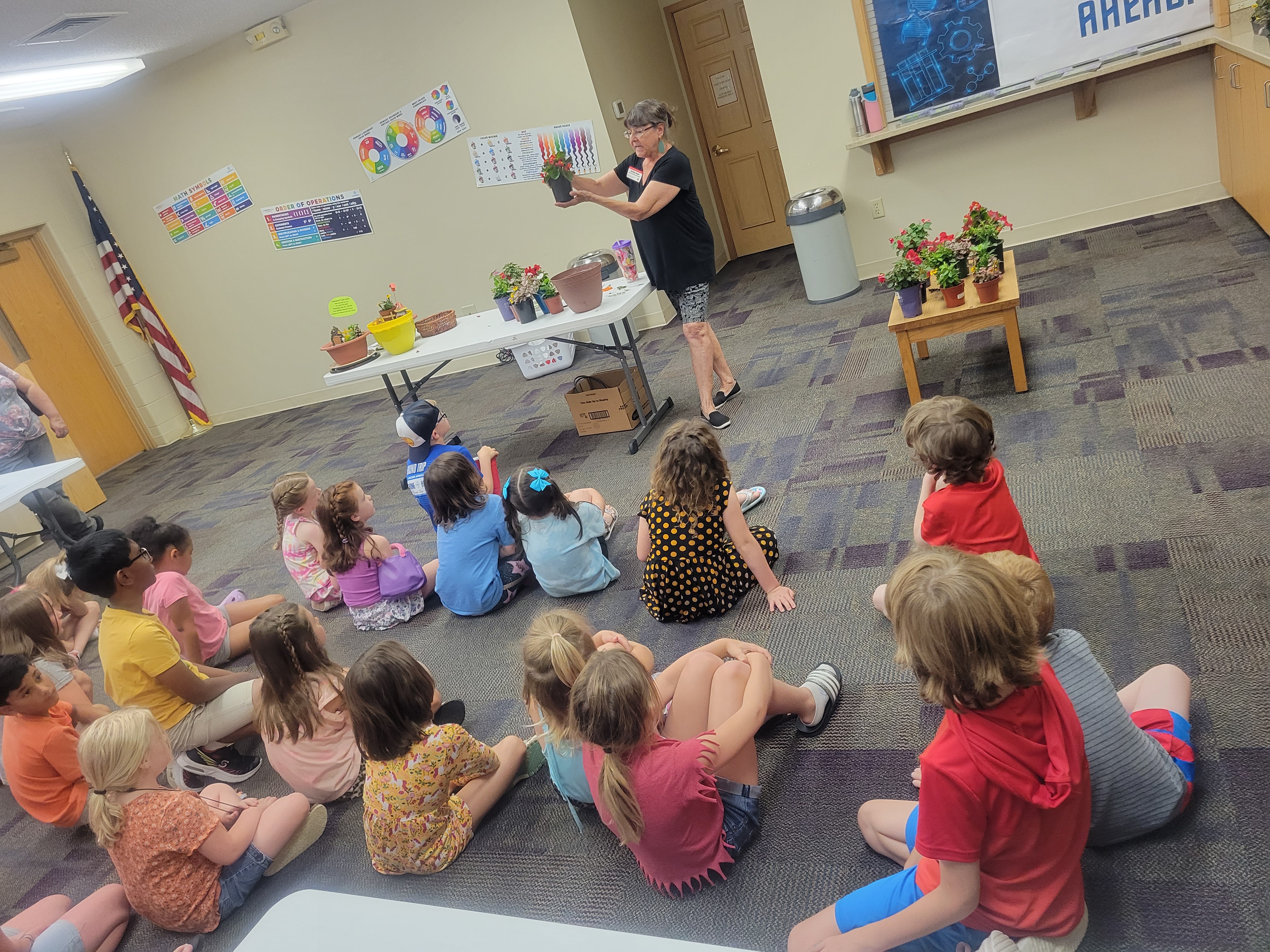 lots of children sitting on the floor listening to a lady talk about the flower in her hand