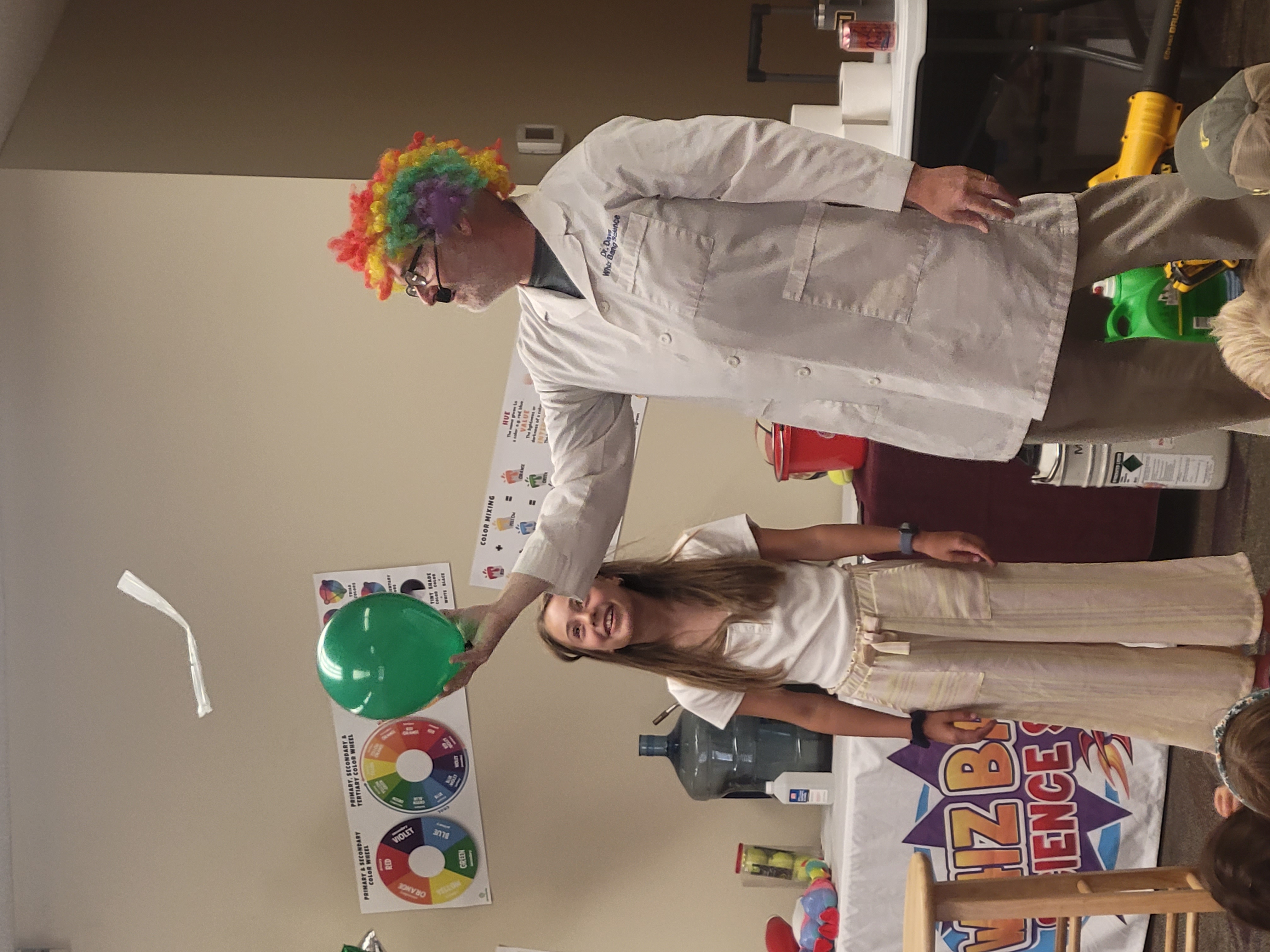 science presenter in a rainbow clown wig using a balloon and static to hold up part of a plastic bag